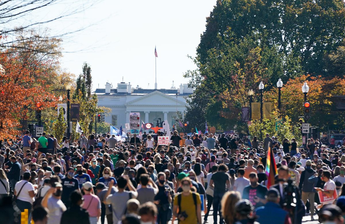 On a road framed by fall trees, the White House stands in the distance. In front of it is a tight mass of people, indistinguishable from one another.