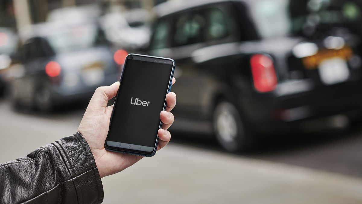 A man wearing a leather jacket holds a smartphone displaying the Uber app.
