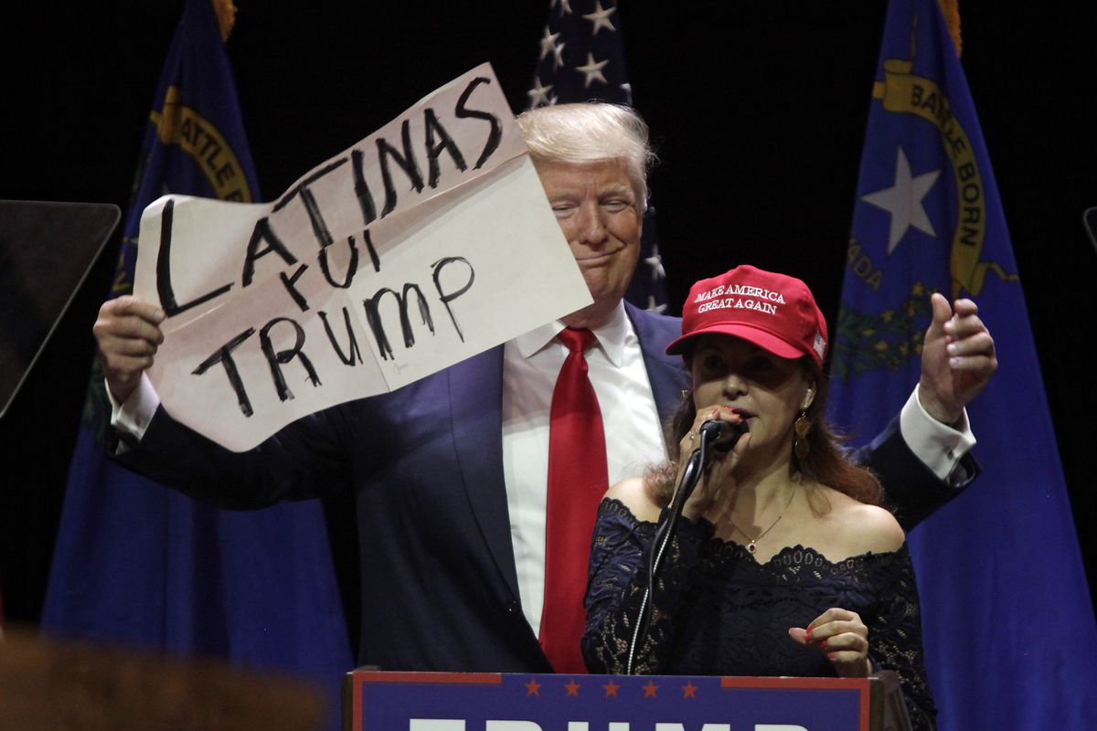 A woman in a MAGA hat speaks onstage while Donald Trump holds a sign behind her that reads “Latinas for Trump.”
