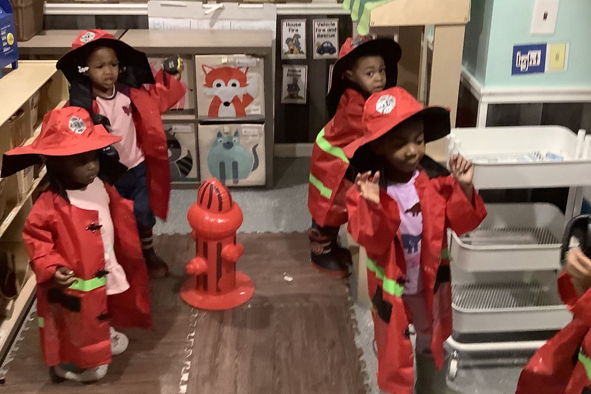 Children wear pretend red fire outfits and helmets next to a pretend red fire hydrant in a preschool classroom