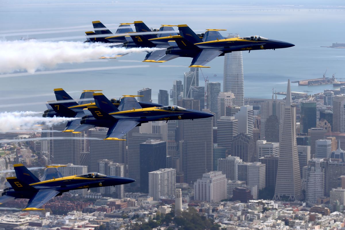 Close-up of blue and yellow fighter jets flying over San Francisco, with tall buildings and the bay below.