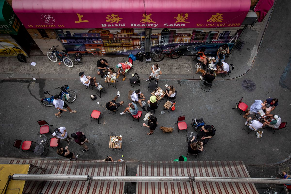 Several tables and chairs set out on a street with people eating at it