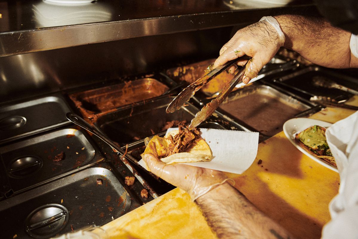 A gloved hand uses tongs to place a small quantity of stewed meat into a tortilla