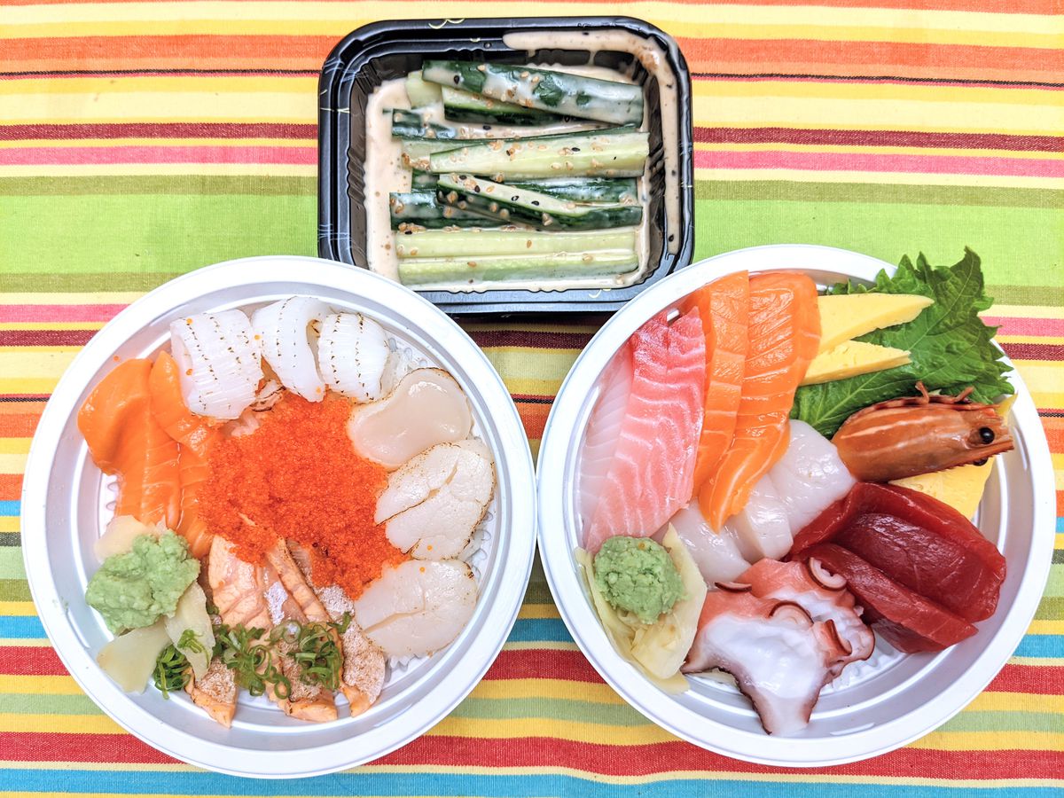 Overhead view of two round white plastic takeout containers filled with a selection of raw seafood and a small black rectangular takeout container with slices of cucumber in a sesame dressing. The containers are on a brightly colored striped tablecloth.