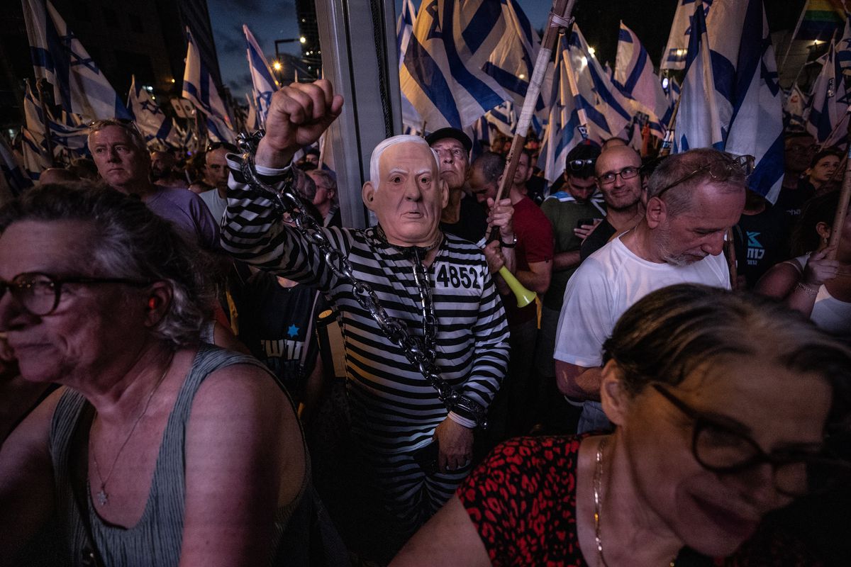 A crowd of protesters, waving blue and white Israeli flags, march through Jerusalem, Israel, on July 23, 2023. One protester wears a black and white striped inmate costume and a rubber mask resembling Netanyahu’s face, and raises a fist in the air.