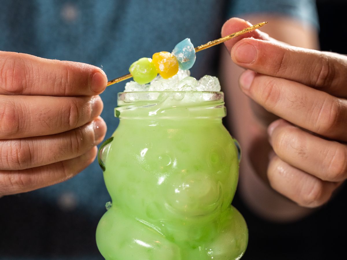 In a glass shaped like a gummy bear, a bartender sets a skewer of mochi treats on top of the green cocktail.