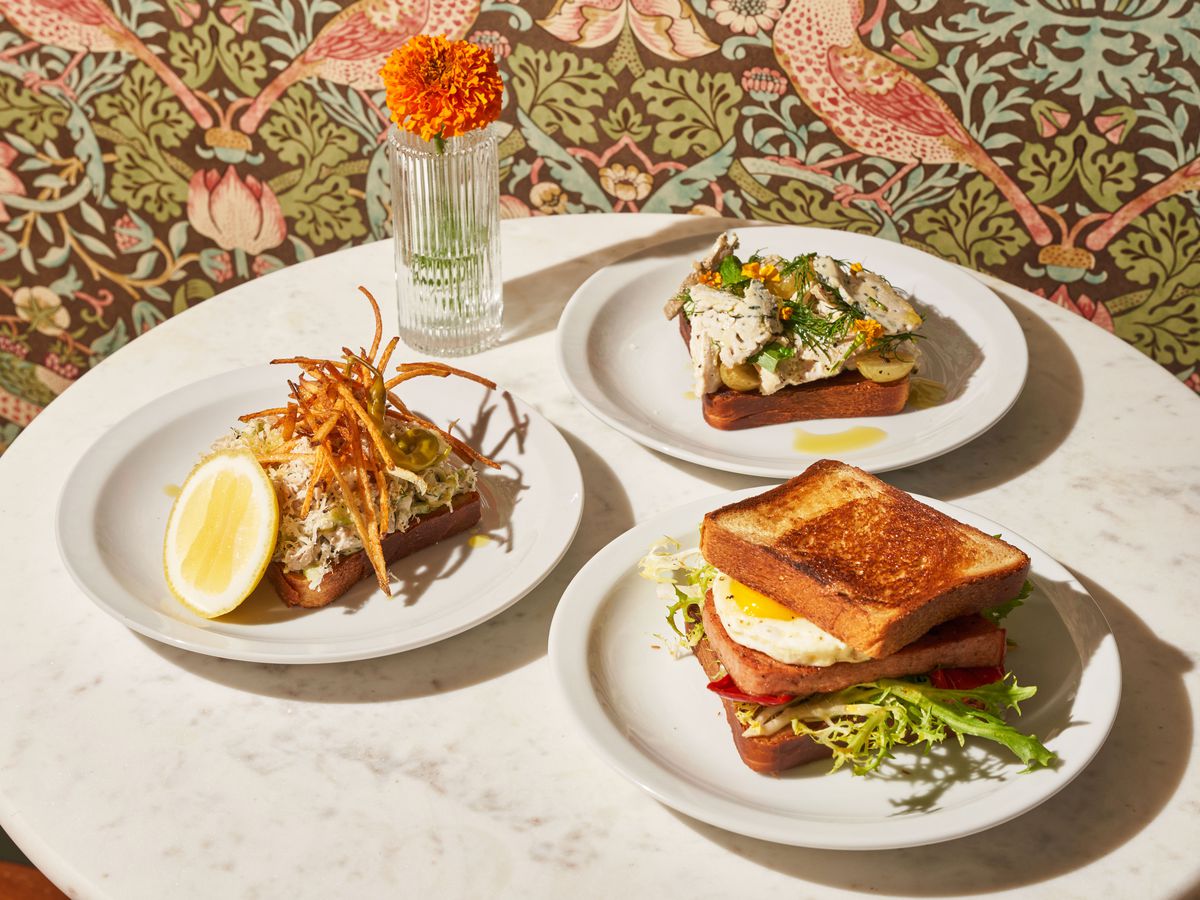 A trio of sanfwiches on white plates sit on a marble table next to a marigold flower in a vase.