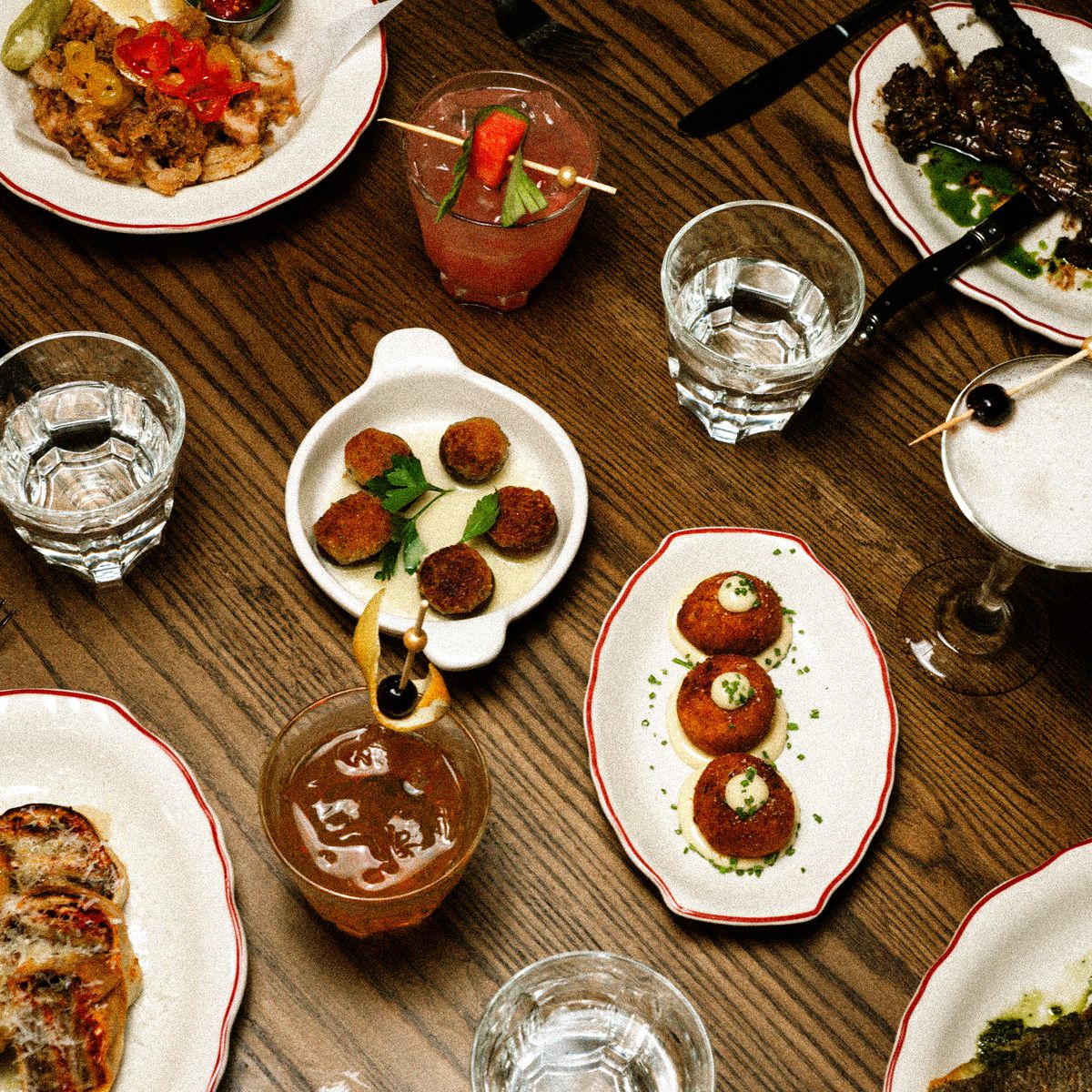 An overhead shot of an assortment of Italian dishes on white plates set on a wood table. Cocktail glasses and cups of waters are scattered throughout. 