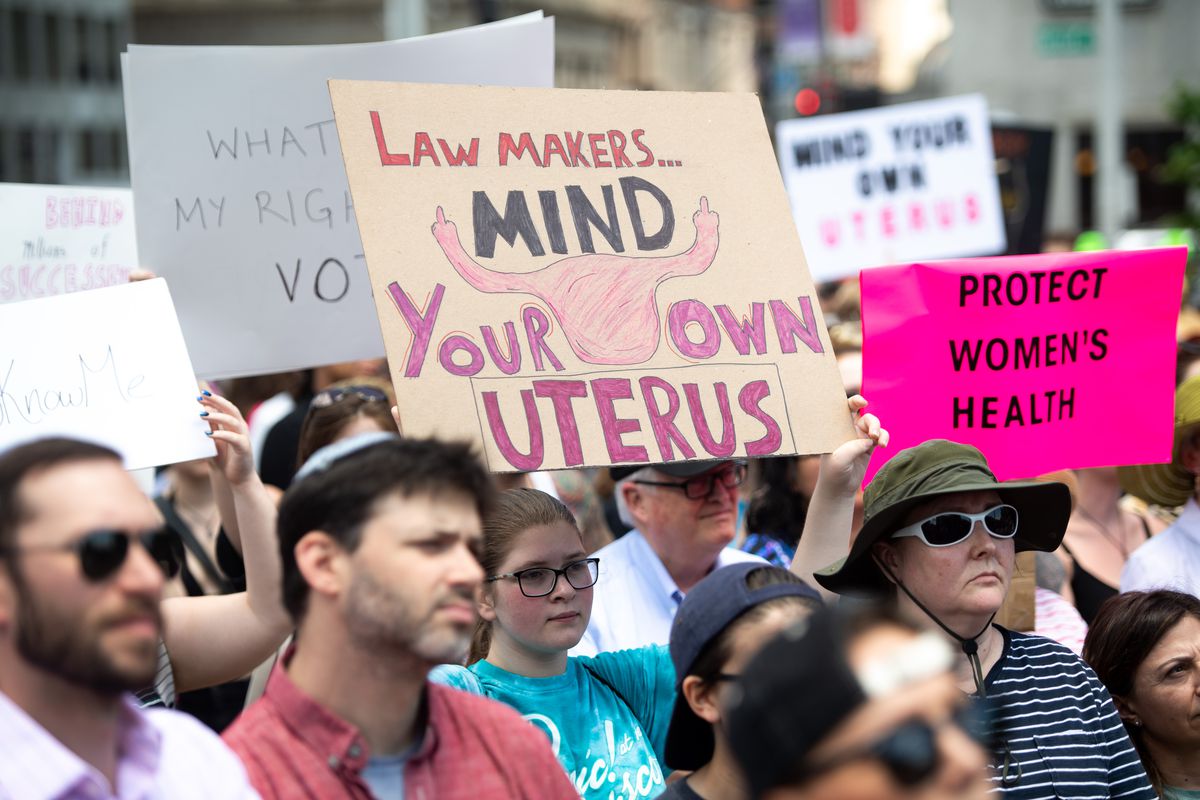 A crowd of protesters, one holding a sign that says, “Lawmakers ... mind your own uterus,” with a drawing of a uterus giving the middle finger. Another protester has a sign reading, “Protect women’s health.”