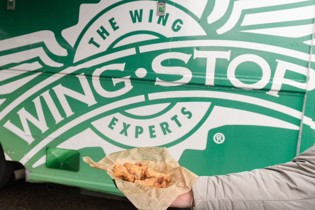 A hand holds out a fry basket filled with fried chicken wings against the Wingstop logo in the background.