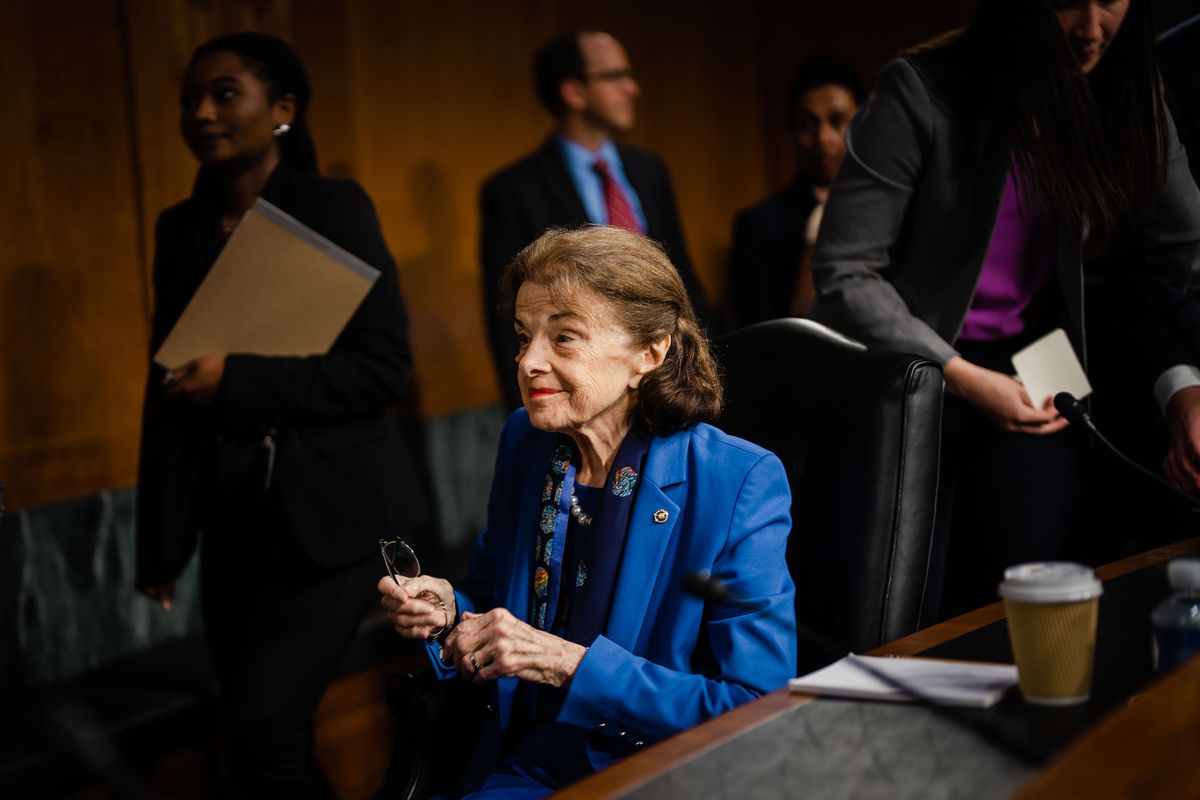 Feinstein, her short brown hair tucked behind her ears, smiles under a committee chamber spotlight. Seated, she wears a royal blue suit and black blouse, her glasses in her hand. She’s turned to the side, and looking upward slightly, as if about to address someone not seen in this image.