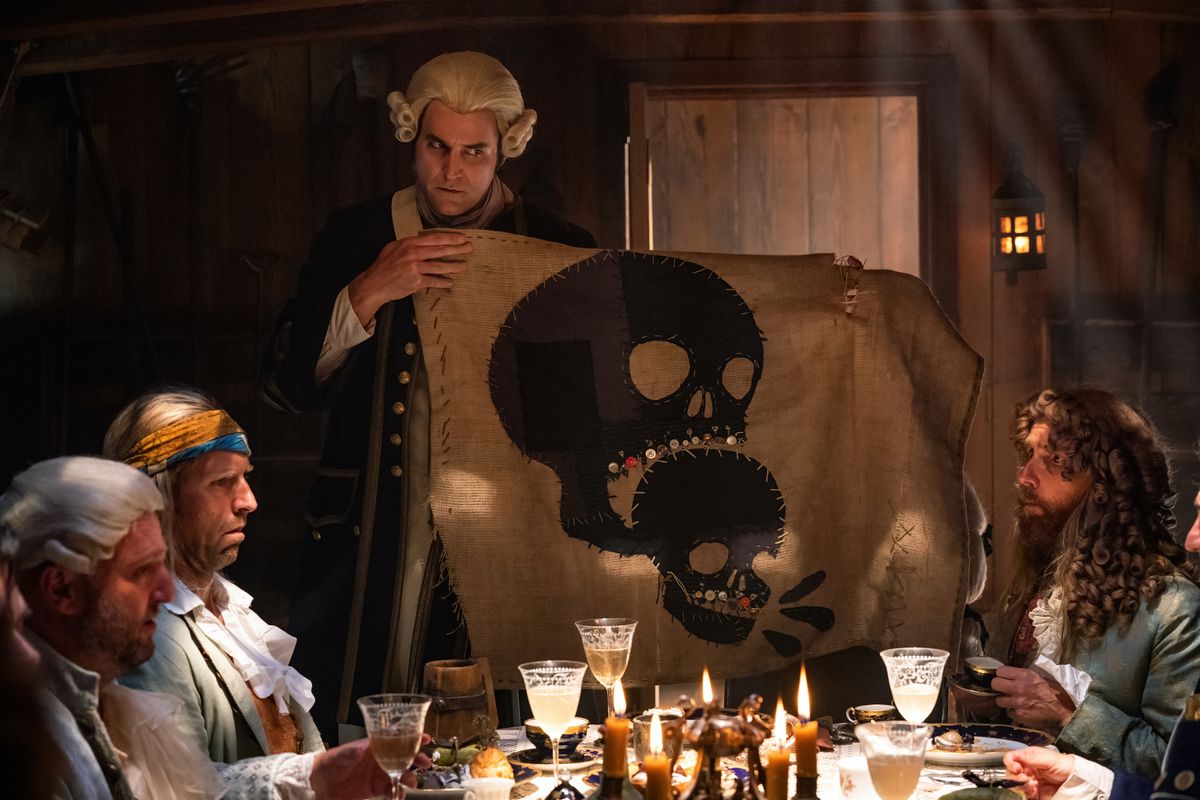 British navy man Connor Barrett holds up a hand-sewn pirate flag with a skull biting a smaller skull, as pirates Ewen Bremner, Matthew Maher and Nat Faxon, disguised as gentleman sailor nasty, very attractive in Our Flag Means Death