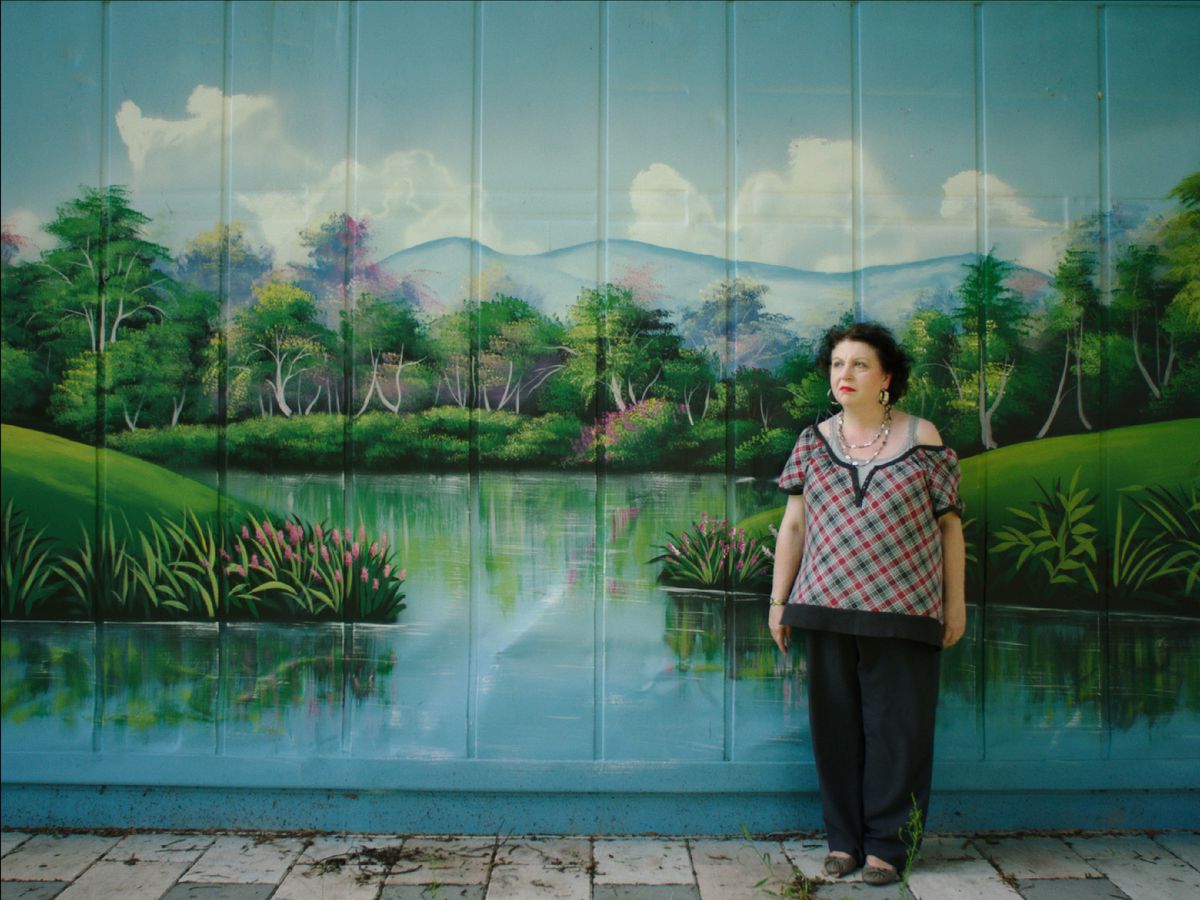 A woman stands in front of a giant painted mural depicting a lush green scene.