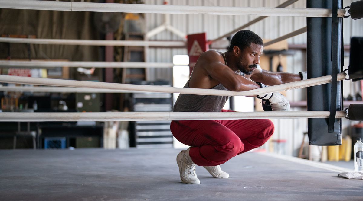 Adonis Creed (Michael B. Jordan), in sweats and white boxing gloves, crouches in the corner of a gym boxing ring and looks grimly determined in Creed III