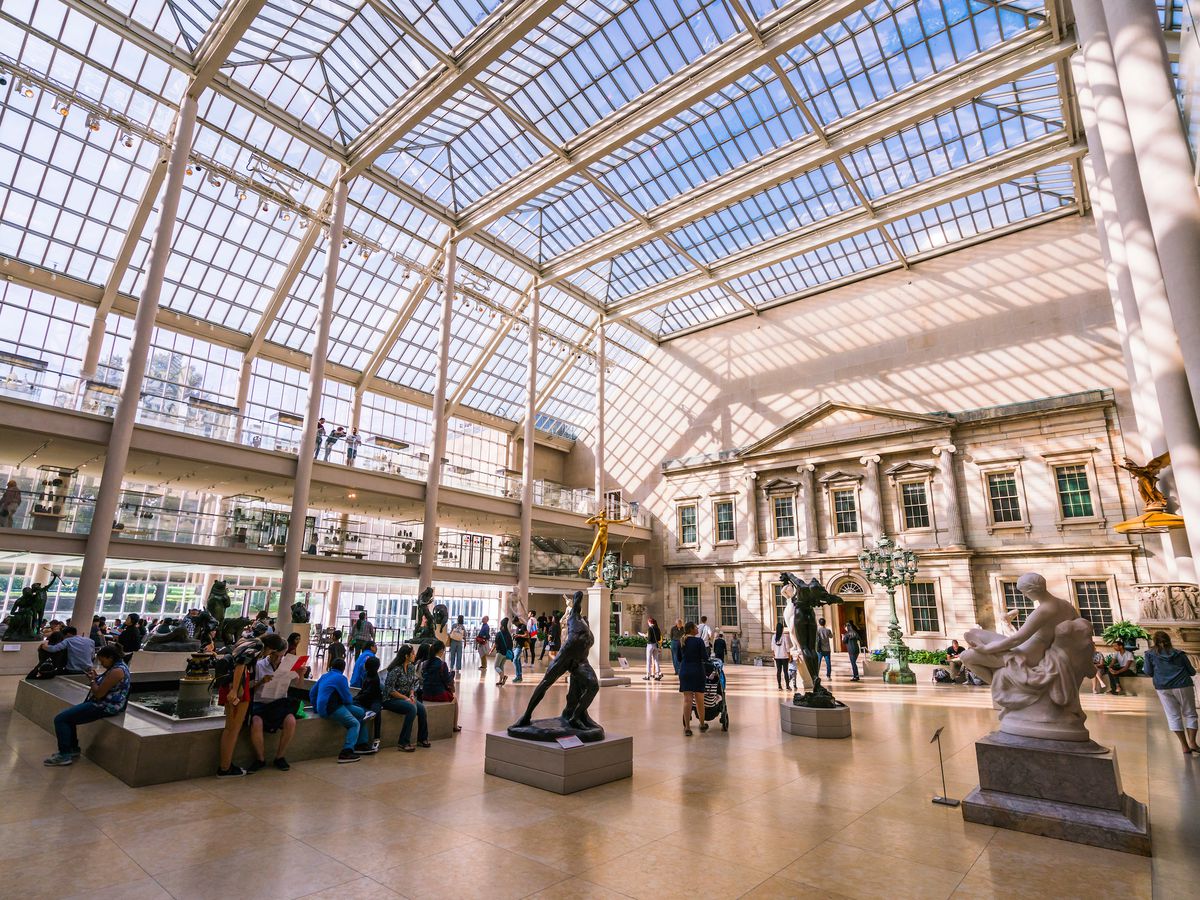 The interior of a museum. Some of the walls are glass. There are statues and sculptures. People are sitting and standing in the room looking at the various art objects.