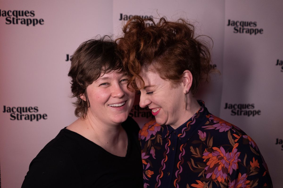 Two women smile and hug each other in front of a Jacques Strappe backdrop.