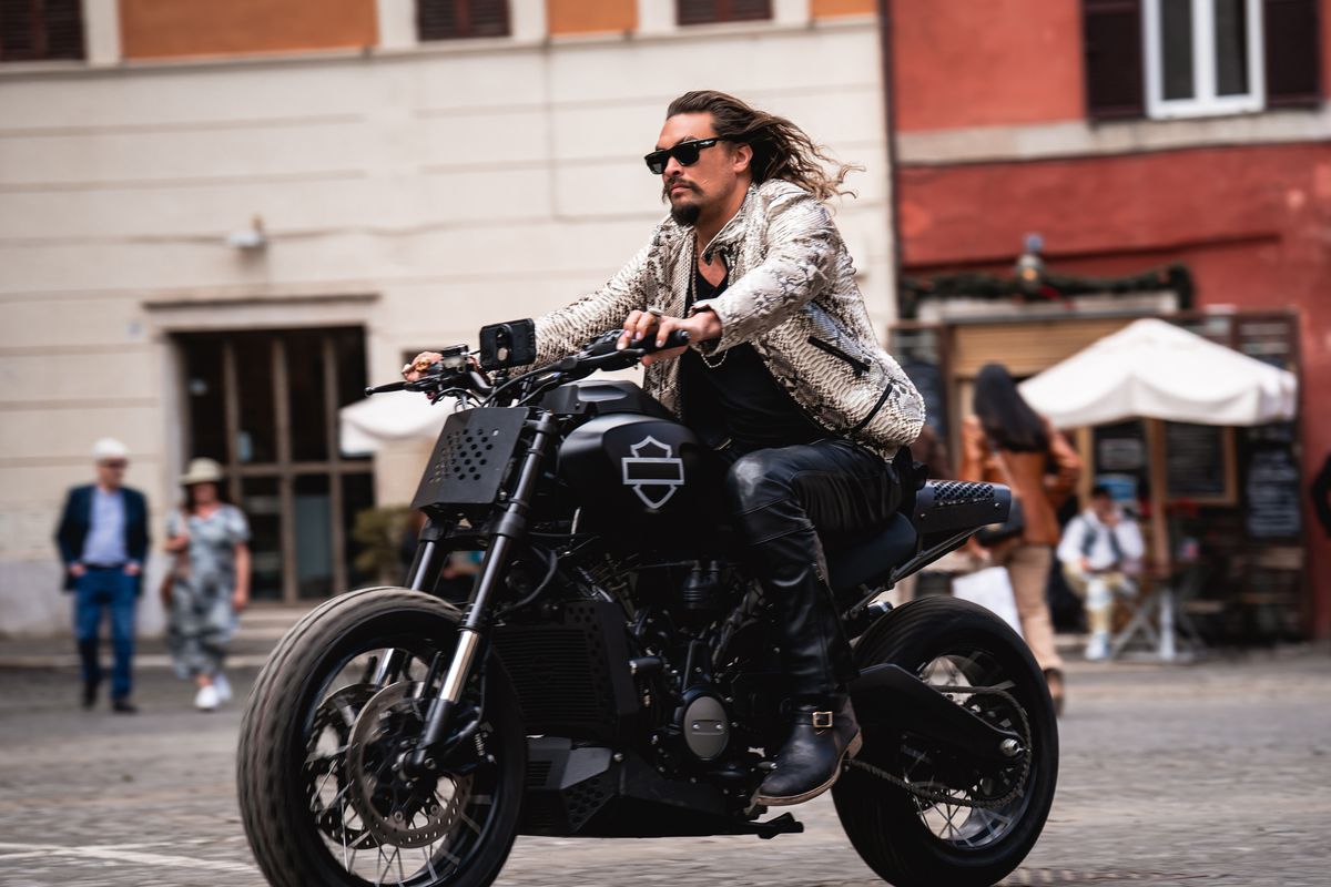 A large man with long hair riding a large motorcycle through a European street.