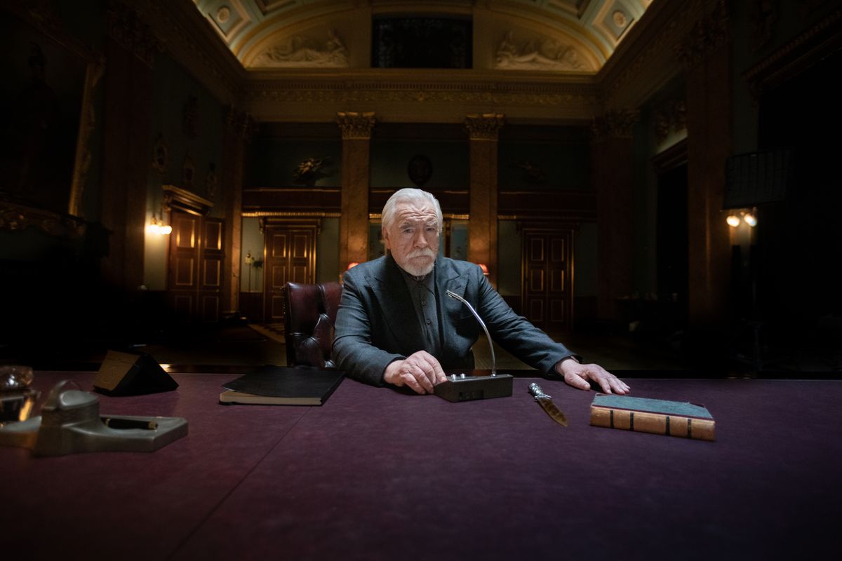 Brian Cox in a dark suit, seated at a table in an opulent room, with an intercom, a knife, and a book before him