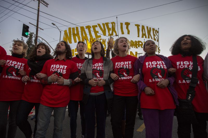 Eight people locking arms outdoors, each wearing a red t-shirt that reads “Fight for $15 and a Union.” 