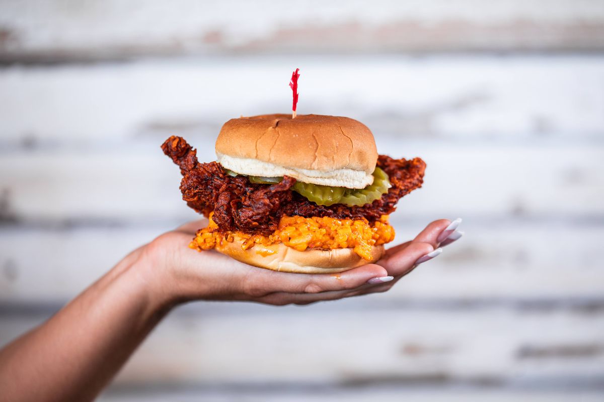 A hand holds a fried chicken sandwich on a bun with a thick layer of chili cheese on the bottom. 