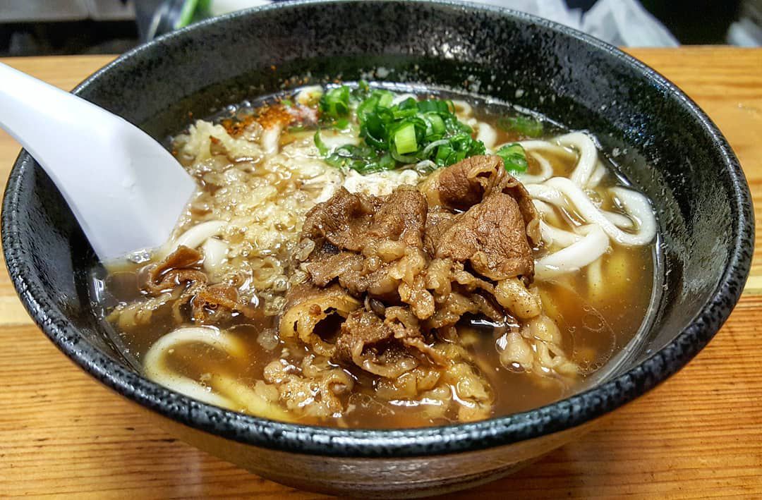A deep black bowl is full of udon soup in a thin brown broth, topped with thinly sliced beef.