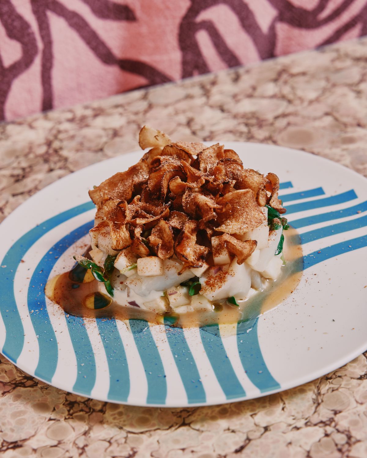 A circular pile of raw seafood topped by crispy brown chips on a white plate decorated with wavy blue stripes.