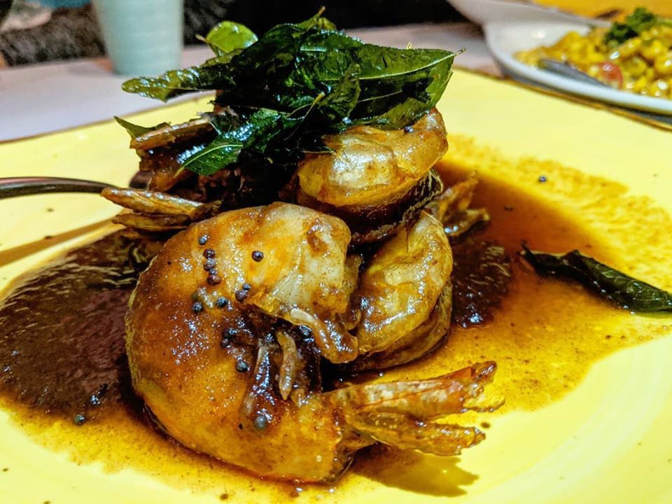 Several shrimps in a dark brown sauce sit on a yellow plate, garnished with fried herbs