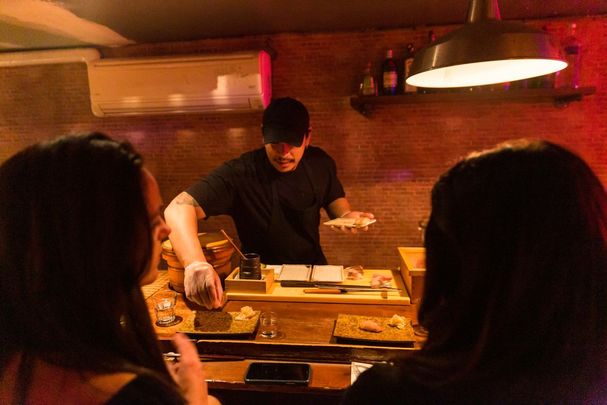 A sushi chef serves nigiri sushi to two patrons at the bar