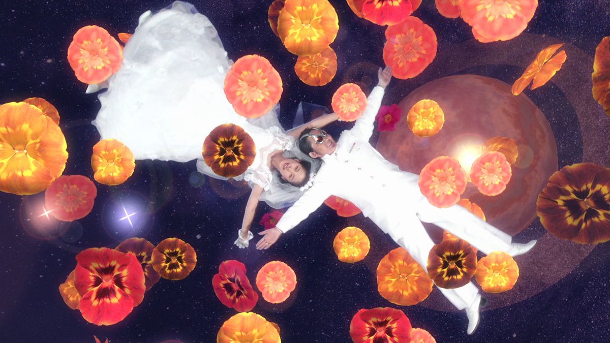 A Japanese woman in a white bridal gown and tiara and a Japanese man in a white military-esque suit and silver goggles with red lenses lie on their backs, arms spread, smiling, against a backdrop of space with a large ringed planet behind them, and huge multicolored pansy blossoms spangling the image in Takashi Miike’s The Happiness of the Katakuris