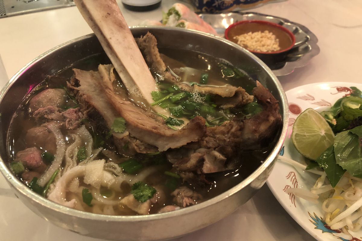 A bowl of pho in a metal bowl topped with a whole beef rib.