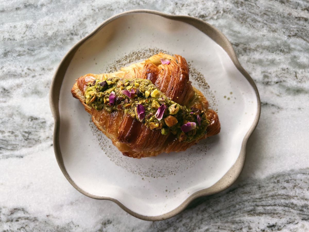 A croissant decorated with rose and pistachio crumbles sits on a stylish plate on a marble counter.