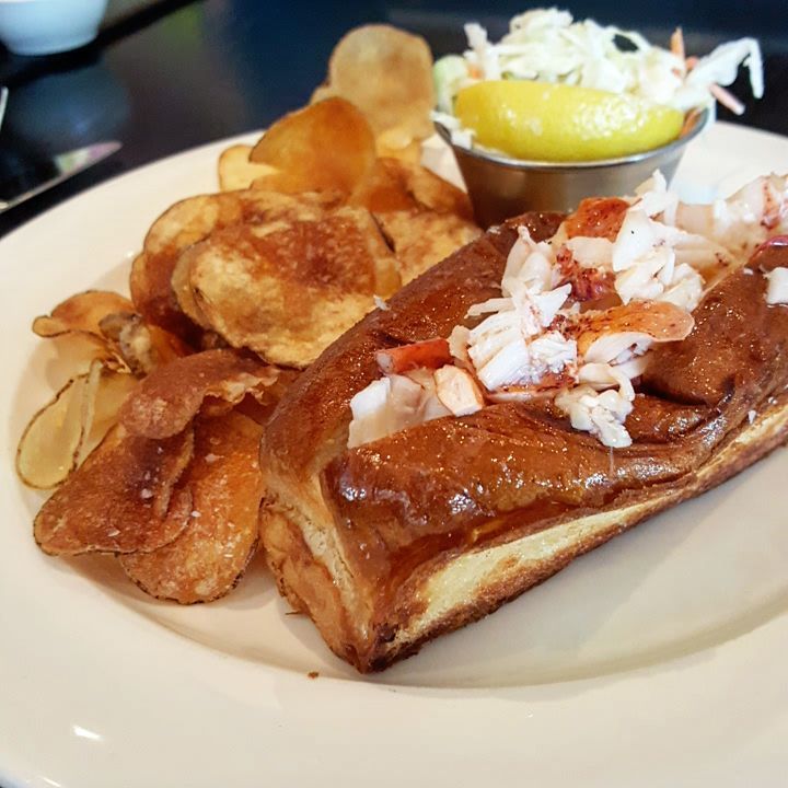 Lobster is served on a buttered and griddled hot dog bun, and is accompanied by chips, slaw, and a slice of lemon.