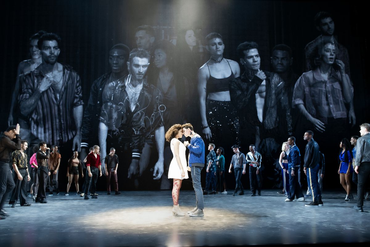 A man and woman embrace at the center of a stage, under a spotlight. Behind them, a crowd lurks in a circle, and above the crowd are larger-than-life video projections of the cast.