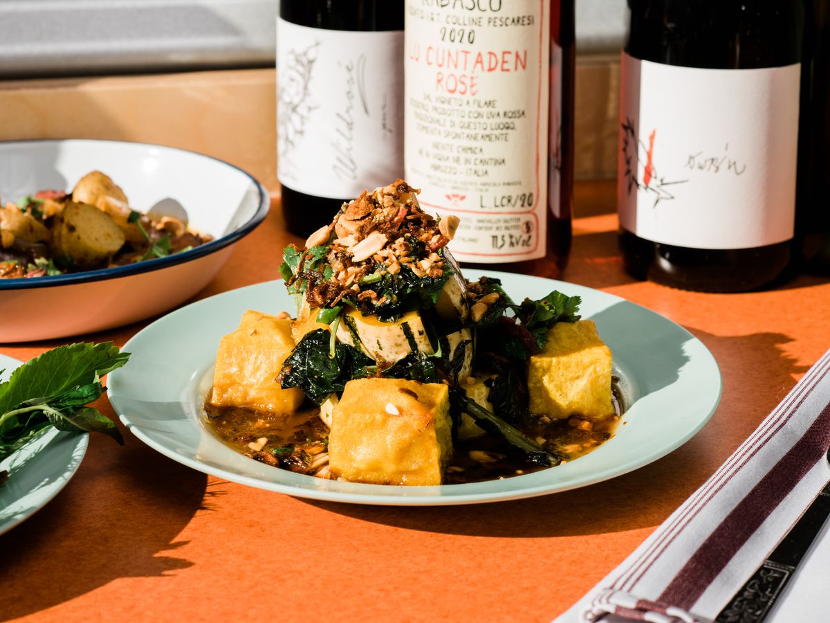 Large cubes of tofu surrounding a pile of greens and peanuts, served beside bottles of wine. 