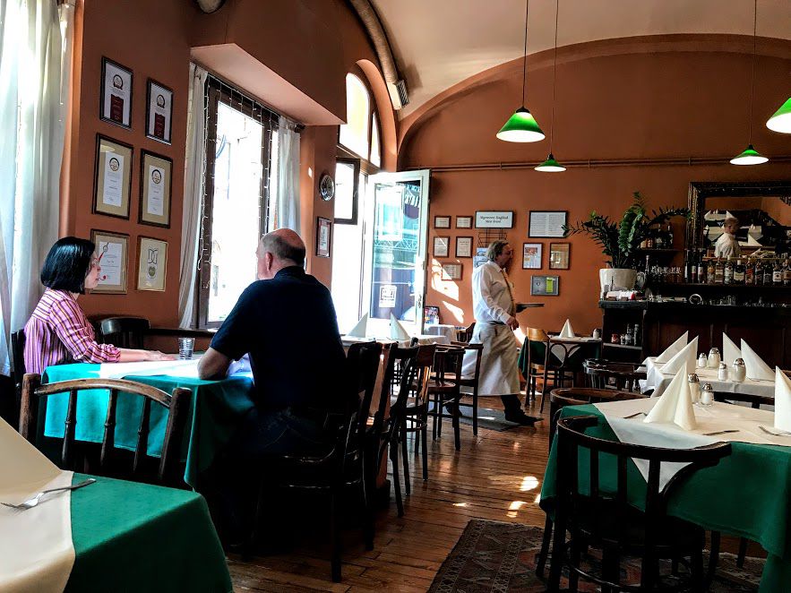 A cafe where a few patrons dine at a table and someone comes through the door. There are photos on the walls and green tablecloths on the tables. 