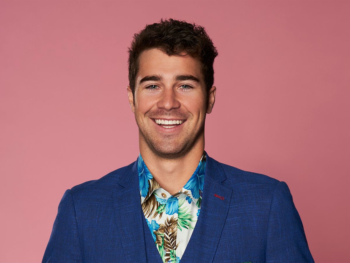 A white man with curly hair, Cole Barnett from season 3 of ‘Love Is Blind’ on Netflix, is pictured on a pink background in a blue three-piece suit with a Hawaiian shirt.