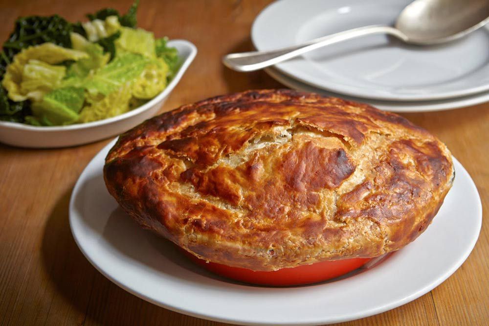 A beef pie in a ceramic dish, with buttered greens on a plate in the background
