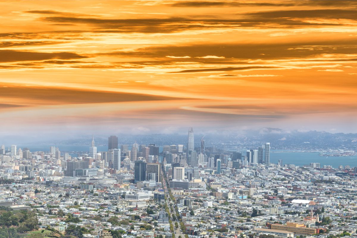 A panoramic, aerial view of buildings in San Francisco, with a layer of gray fog hovering over it and a bright orange sunset coloring the sky above that.