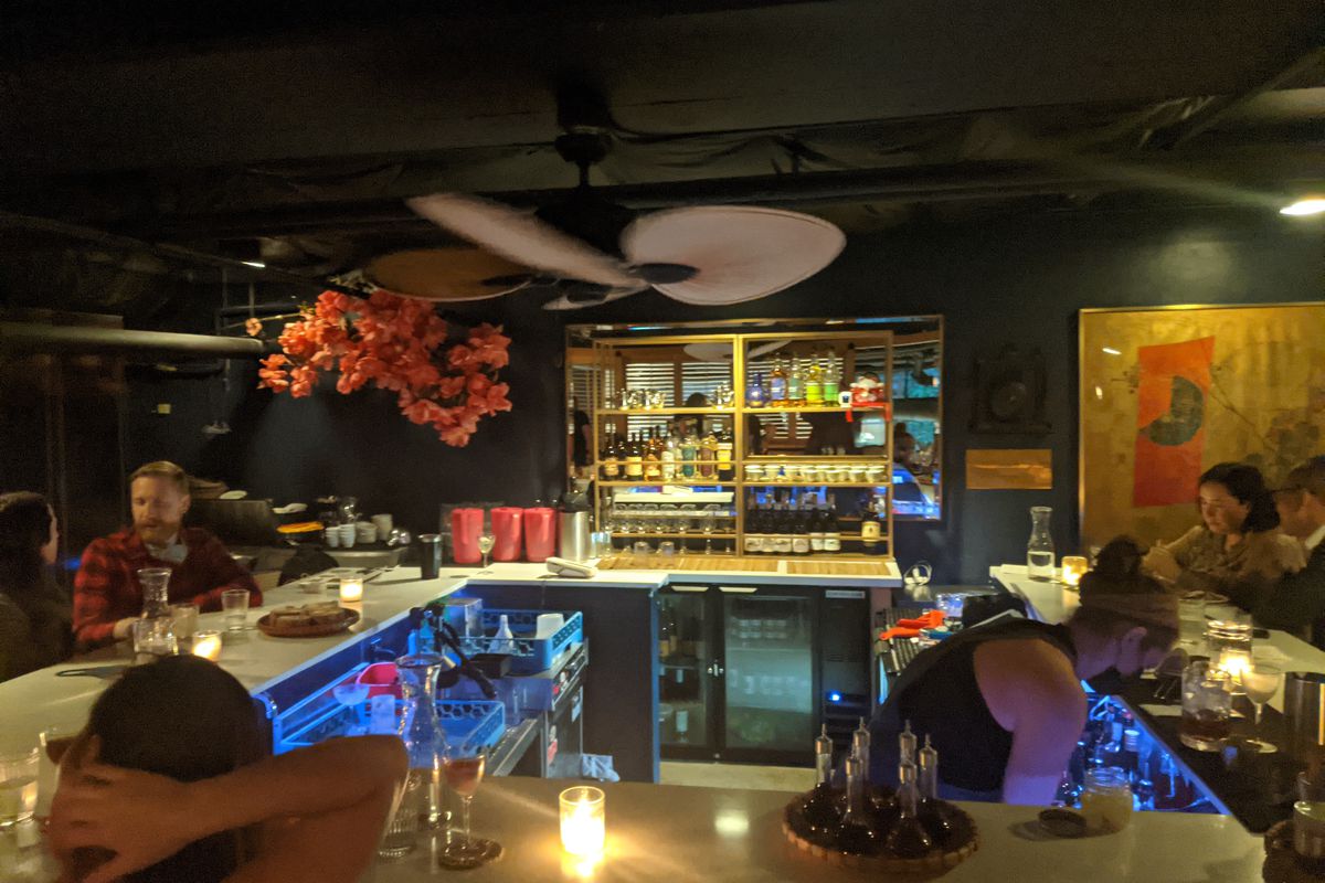 A square-shaped bar with white counters and candles. There are bright orange blossoms in the corner of the dimly-lit room.