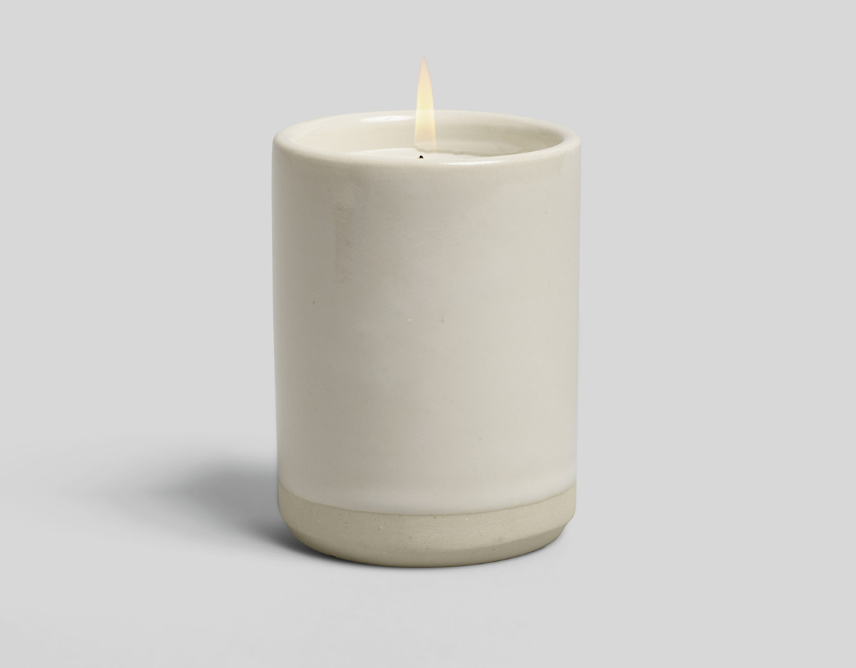 A cream colored sculpted stoneware Idyllwild candle holder. The candle within the candle holder is lit.