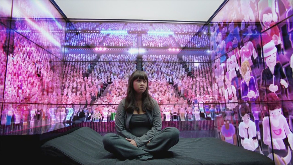 A woman sits in a room surrounded by walls of computer avatars seated in a large auditorium.