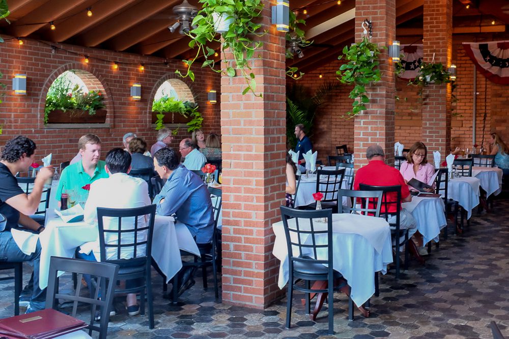 People sit outside at patio tables with white tablecloths surrounded by brick and plants. 