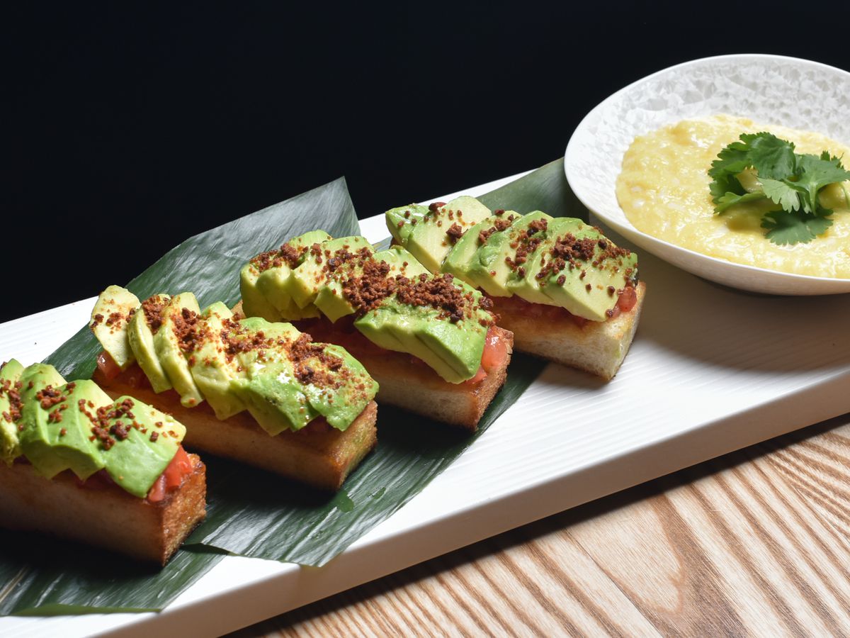 Four perfectly-shaped rectangles of toast topped with thin, green slices of avocado and a dark dusting of dry miso. A bowl of scrambled eggs is also on the plate.