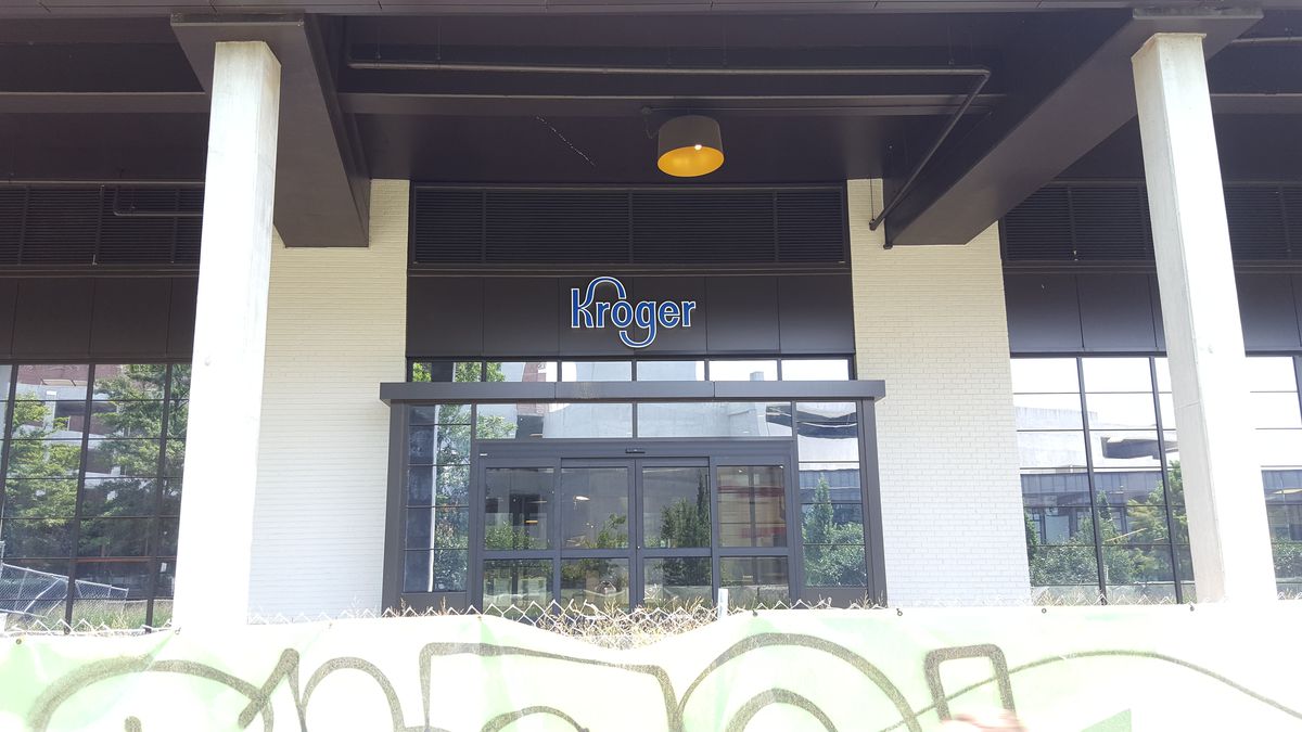 A new Kroger sign is seen behind a construction fence covered in graffiti.