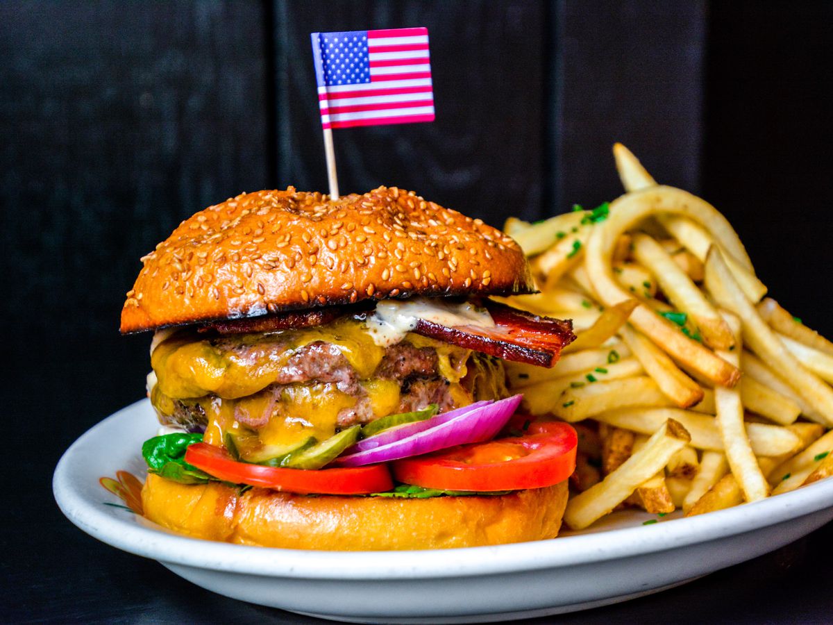 A tall cheeseburger with two patties, tomato slices, pickles, red onions, lettuce, and bacon on a sesame seed bun with an American flag toothpick in the top, on a dish next to french fries.