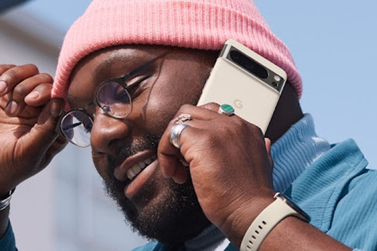 A man in glasses and a knit cap talks on a cellular phone, described by Google as the Pixel 8 Pro in Porcelain.