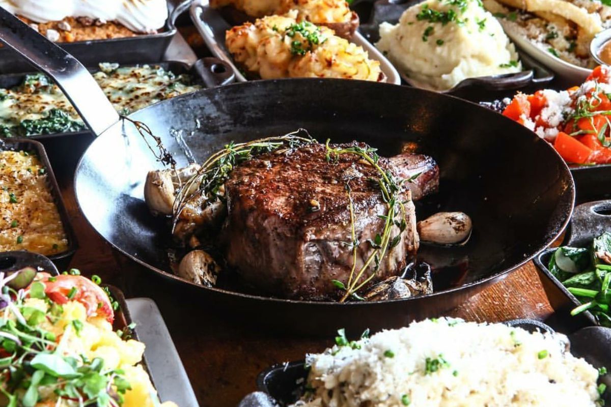 A cast iron skillet with a large steak surrounded by sides of mashed potatoes, rice, and vegetables