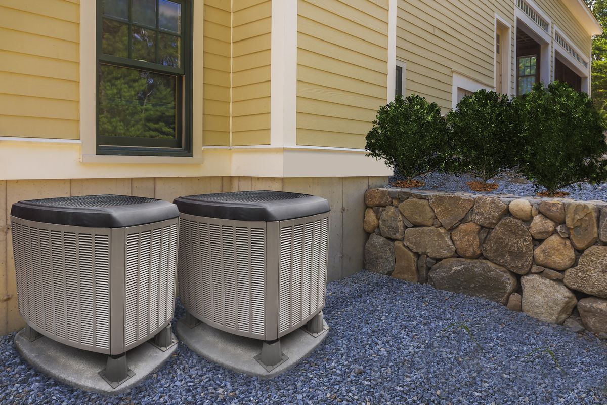 Two heat pumps on the exterior of a yellow home near a stone wall. 