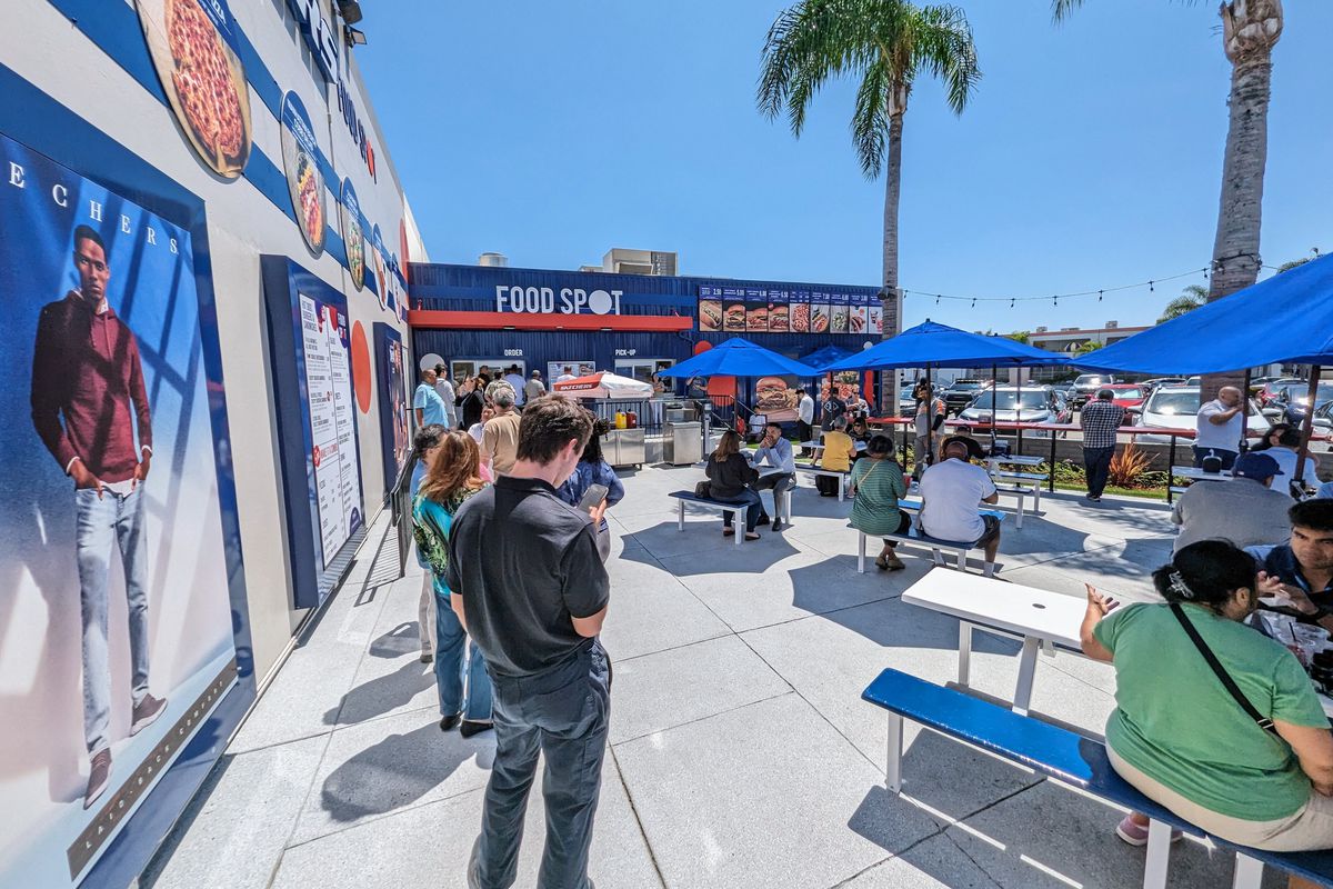 People wait in line at a food window with blue paint accents and outdoor dining area.