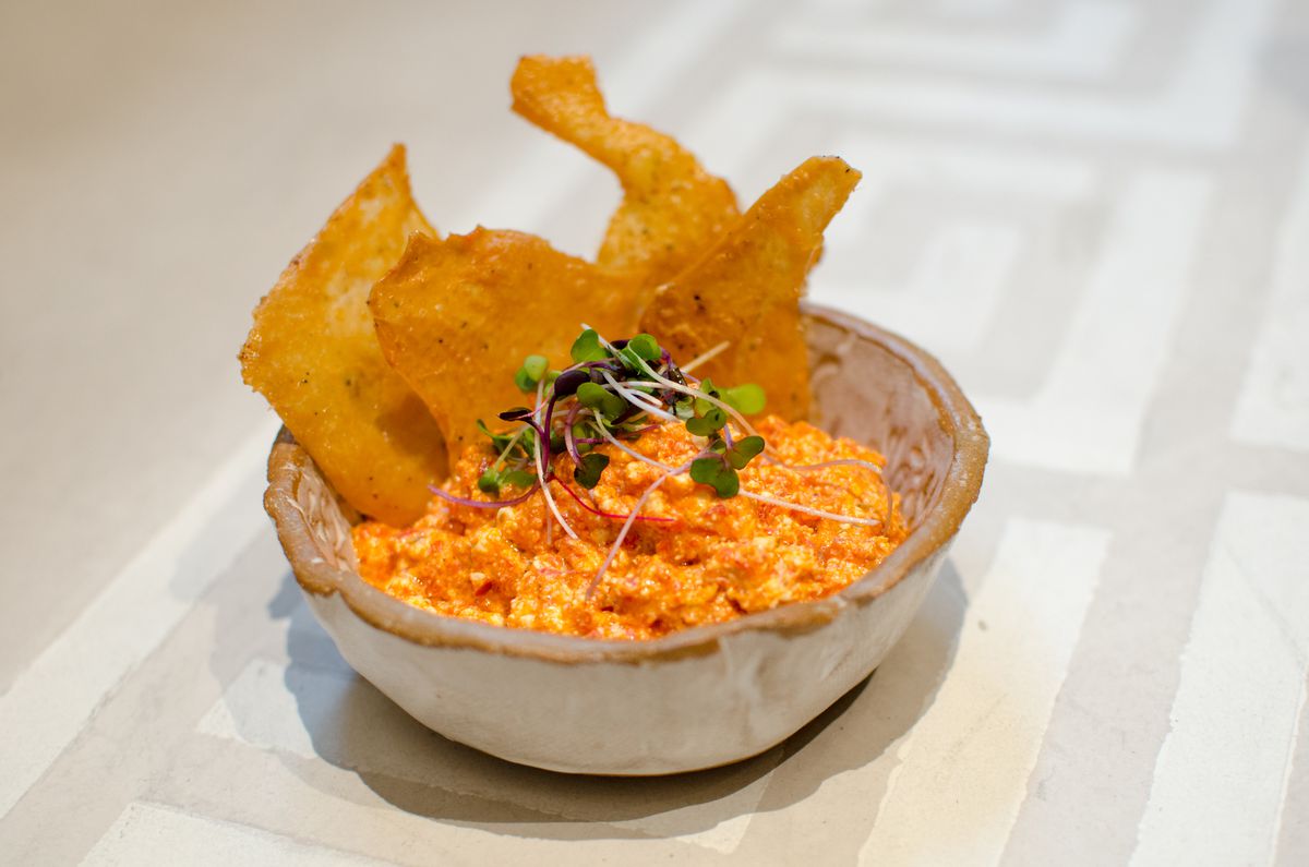 A thick orange dip, garnished with herbs, sits in a rustic stone bowl on a light-colored surface. There are crispy chunks of chicken skin standing up in the dip.